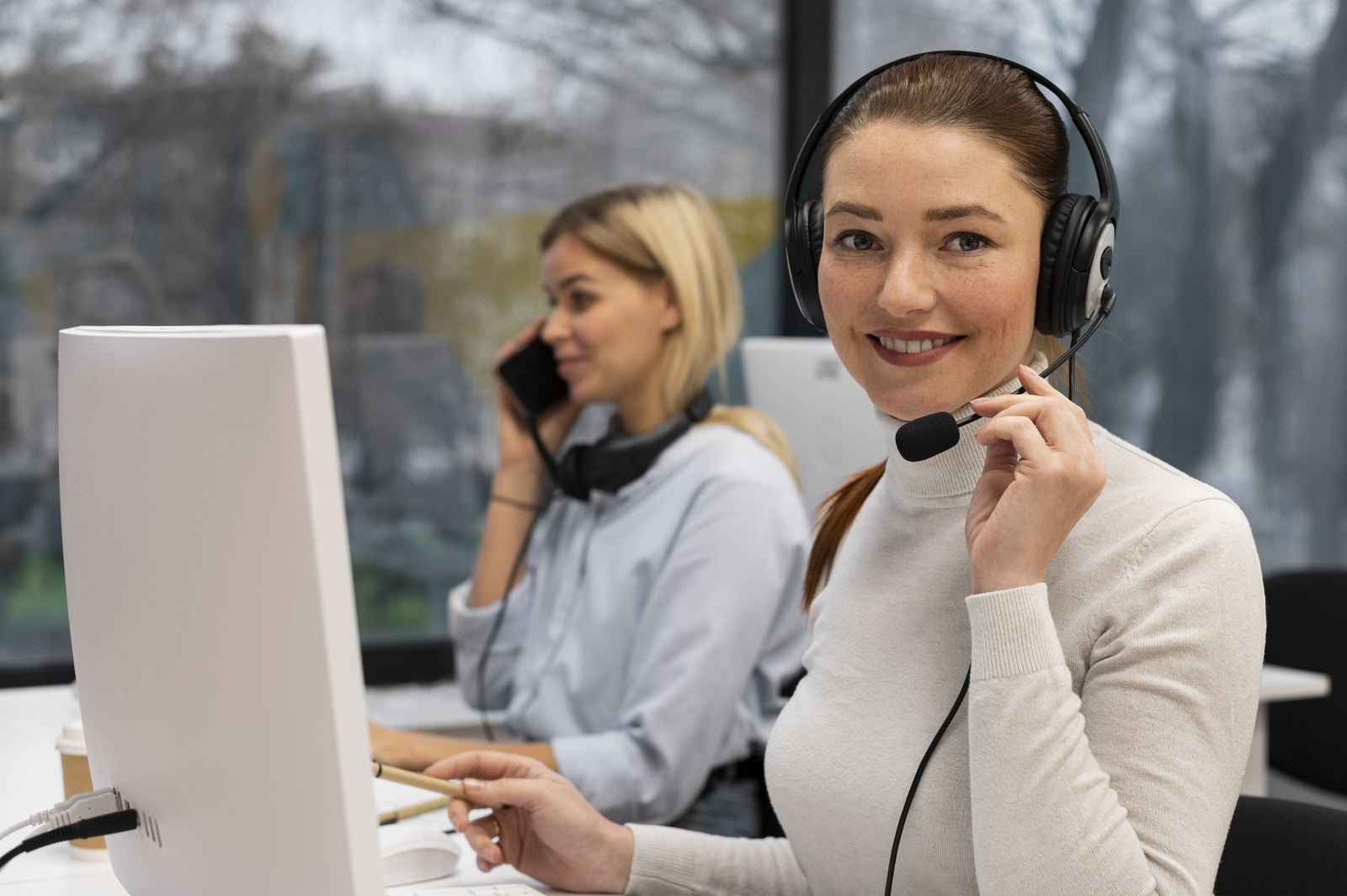 Smiling customer service agent wearing a headset at a desk, with a colleague on the phone behind her