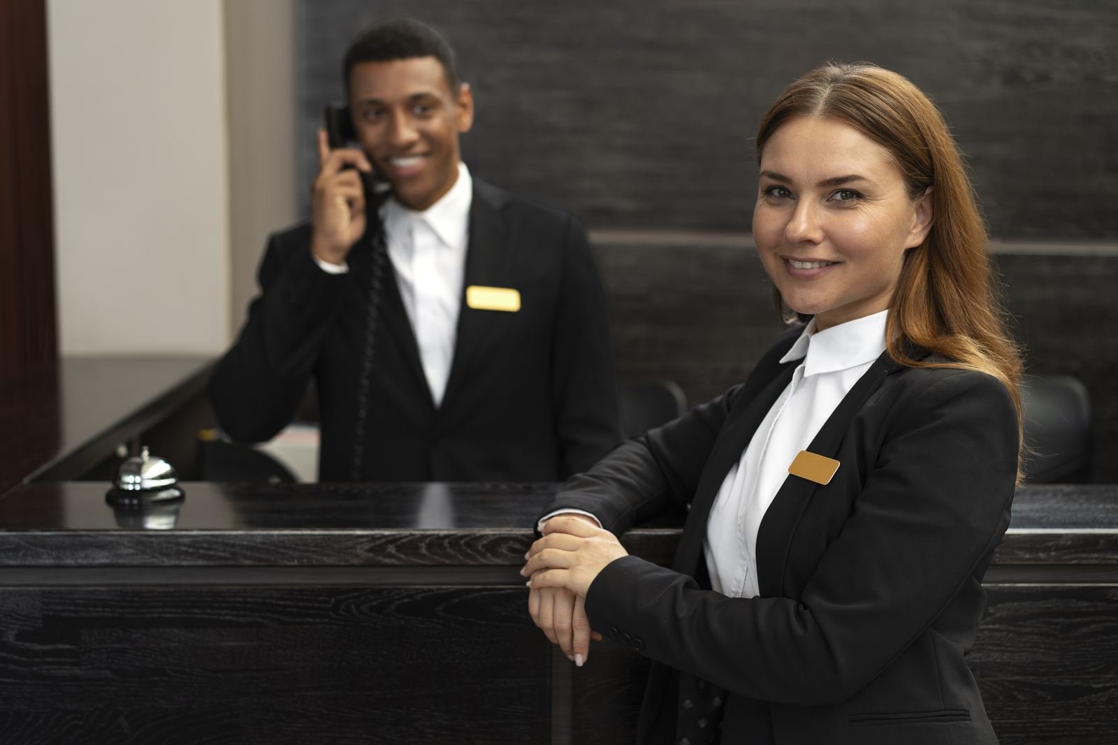Hotel receptionist smiling at the front desk while a colleague takes a phone call