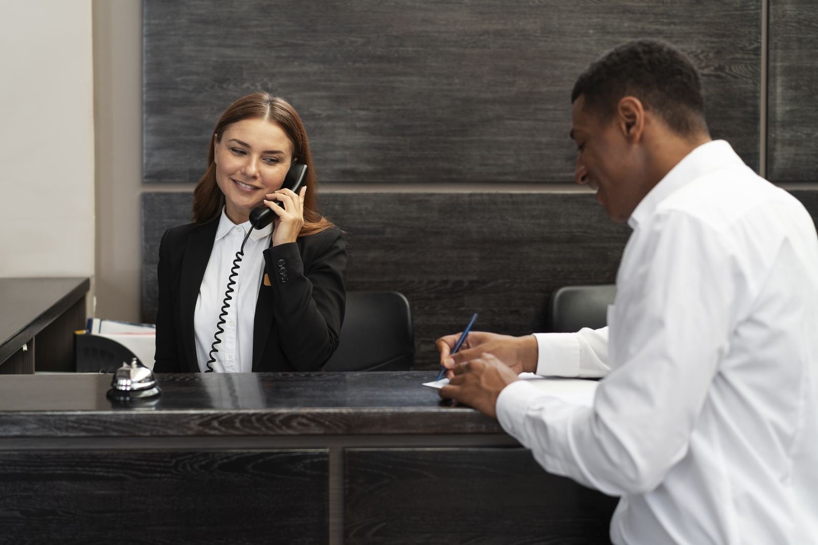 A hotel receptionist on the phone while a guest fills in a form at the front desk