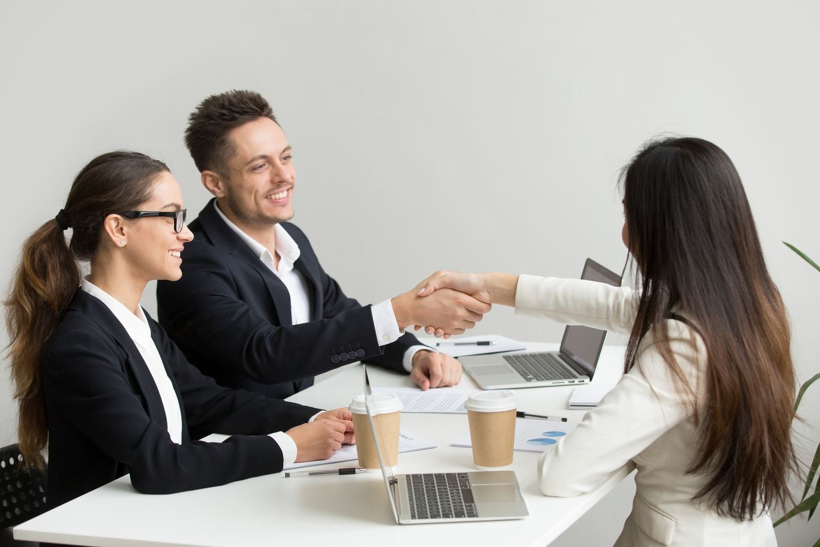Colleagues shaking hands at a meeting table with laptops and coffee cups